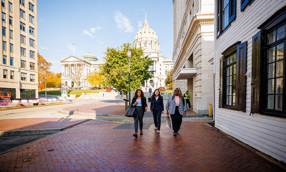 Three students in professional attire walk along a brick sidewalk in downtown Harrisburg on a sunny fall day, with the Pennsylvania State Capitol building visible behind them.