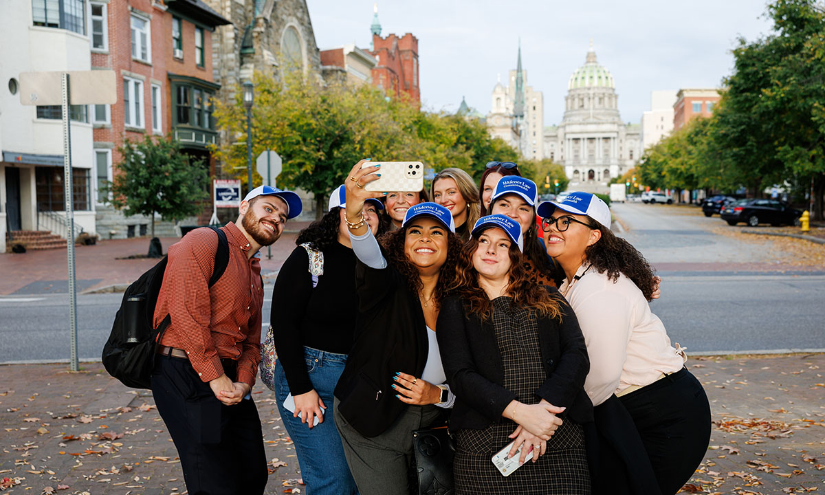 A group of students wearing Widener Law Commonwealth caps takes a selfie on a downtown Harrisburg street in fall, with the Pennsylvania State Capitol dome visible at the end of the block.