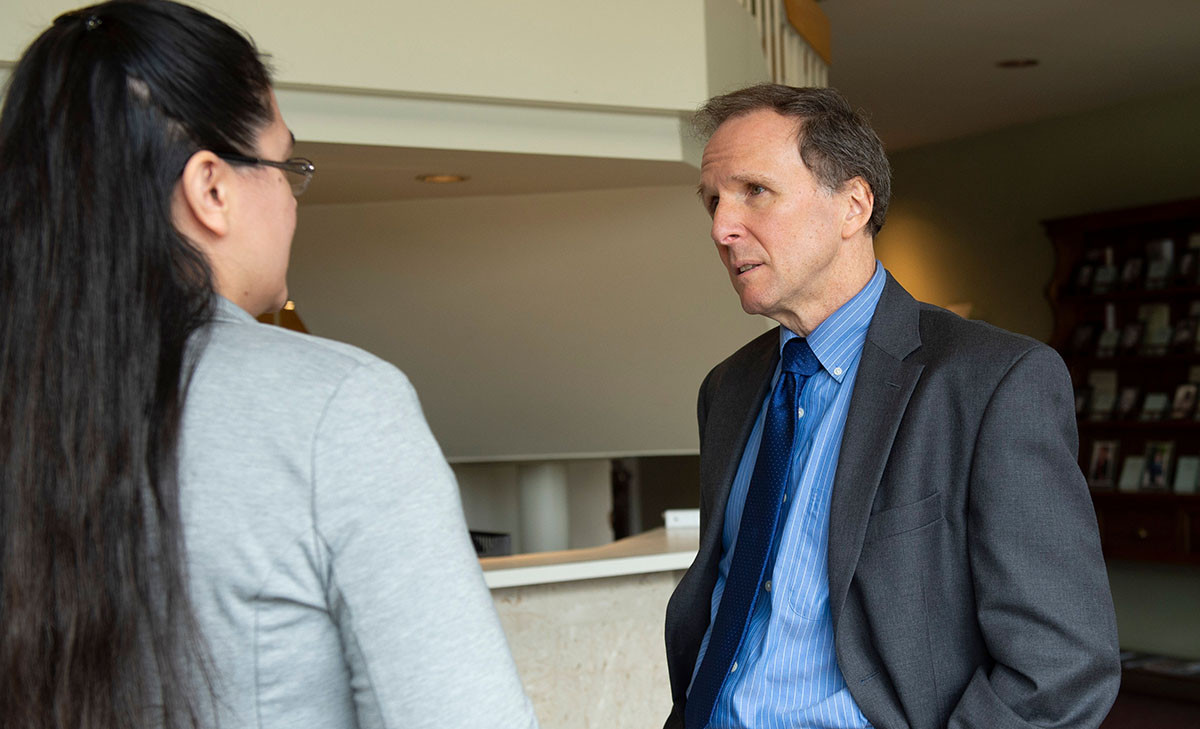 A faculty member in a gray blazer engages in conversation with a student in a campus lobby, with bookshelves visible in the background.