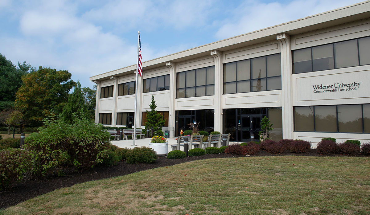 The Widener University Commonwealth Law School building exterior on a partly cloudy fall day, with an American flag out front and benches and planters along the entrance walkway.