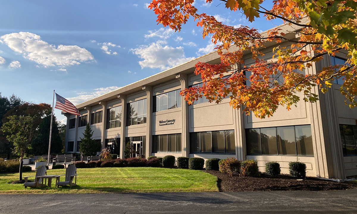 Exterior of the Widener University Commonwealth Law School building on a sunny autumn day, with vibrant orange and red foliage framing the entrance and an American flag flying on the front lawn.