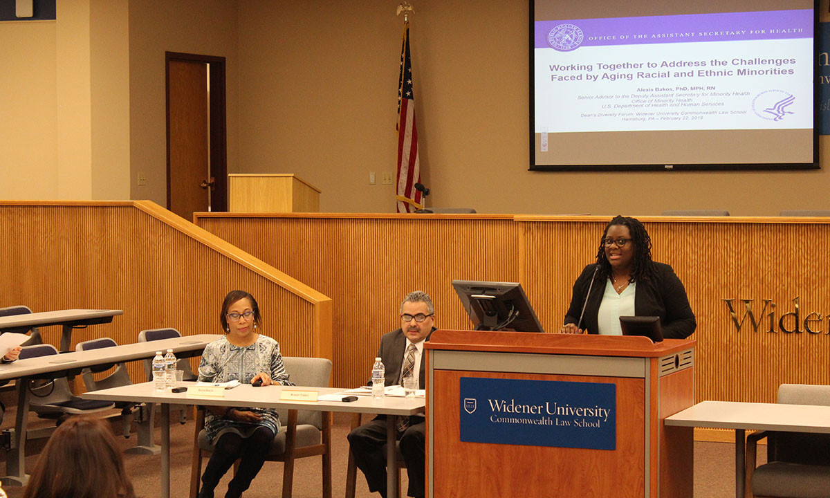 A speaker addresses the audience from a Widener University Commonwealth Law School podium during the 2019 Dean's Diversity Forum, while two panelists are seated at a table nearby. A projected slide behind them is titled "Working Together to Address the Challenges Faced by Aging Racial and Ethnic Minorities," presented by Alexis Bakos, PhD, MPH, RN of the U.S. Department of Health and Human Services.