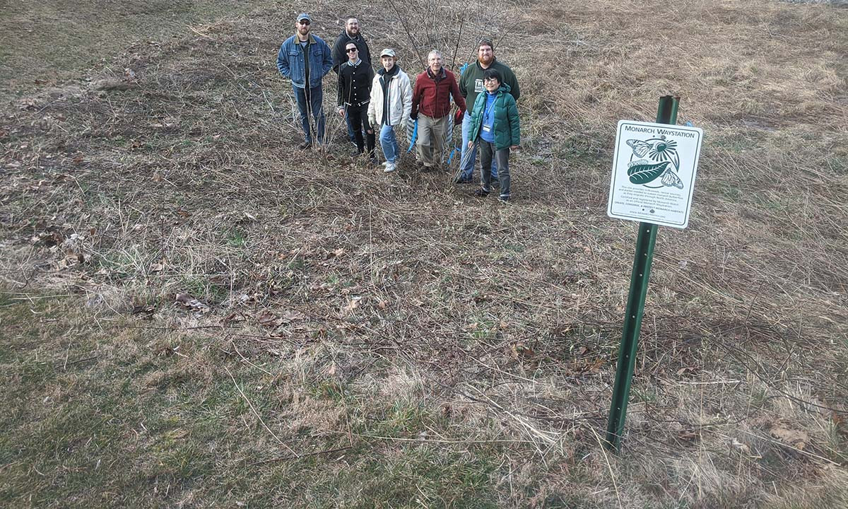 A group of students and community members pose beside a Monarch Waystation sign on a bare winter hillside during an Environmental Law and Sustainability Center habitat restoration project.