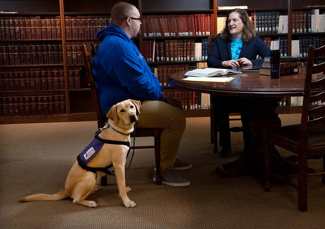 A student seated at a table in a university library speaks with a staff member while a service dog wearing a purple vest sits beside the student.