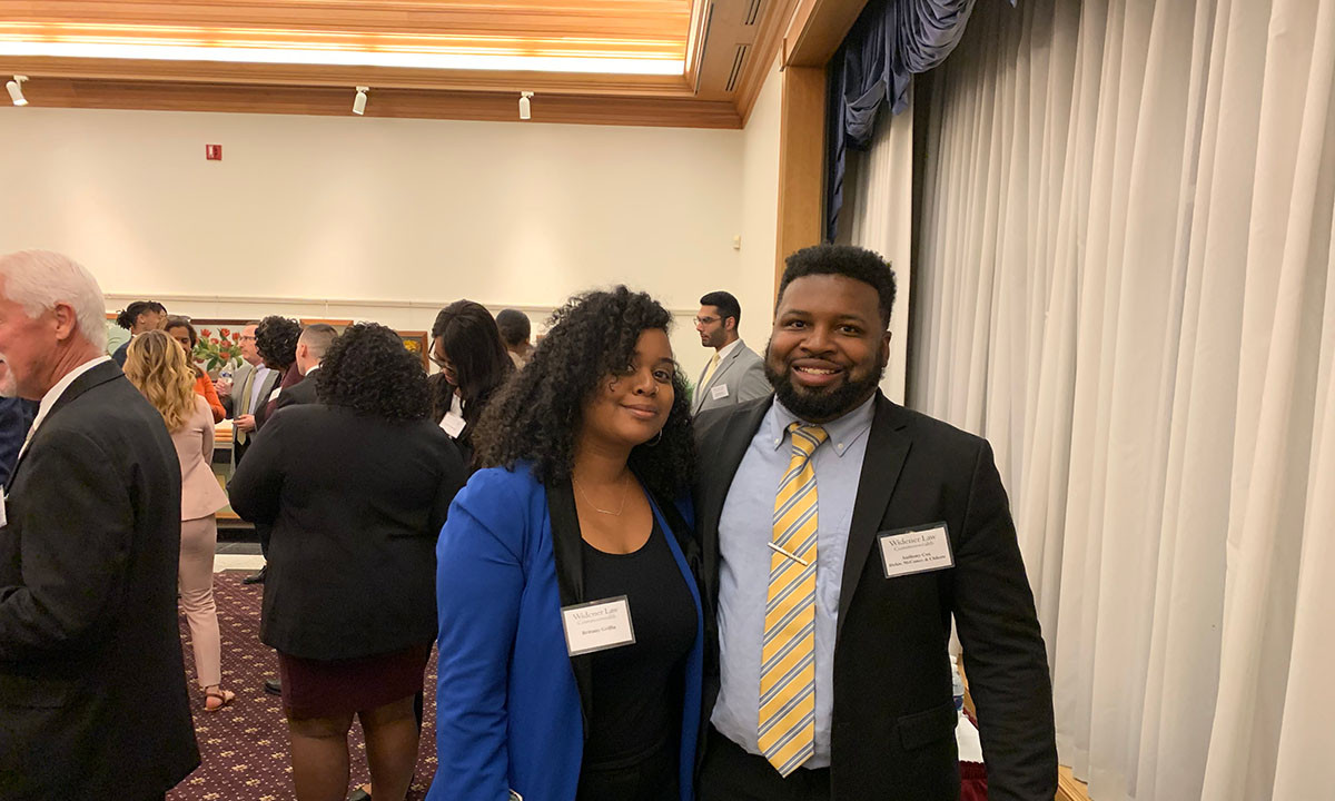 Two students wearing Widener Law Commonwealth name badges pose together at the 15th Annual BLSA Networking Event, with a room full of attendees mingling in the background.