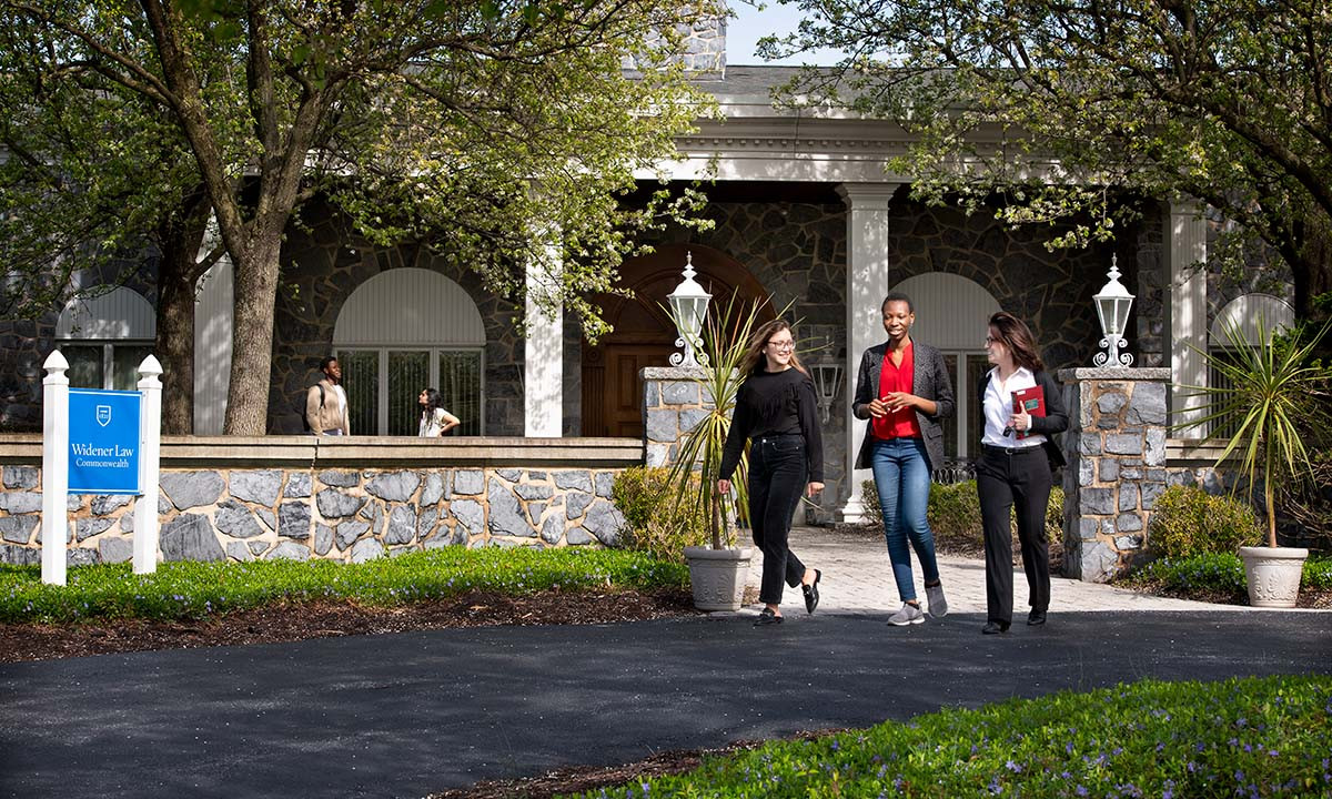 Three students walk together along a path in front of the Widener Law Commonwealth administration building on a sunny spring day, with trees in fresh bloom and a Widener Law Commonwealth sign visible near the entrance.
