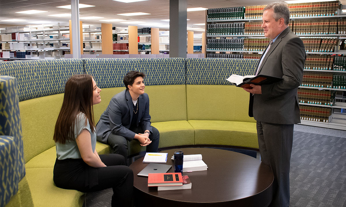 A law librarian consults with two students seated on a curved lounge sofa in the law library, holding an open reference book while law volumes line the shelves behind them.