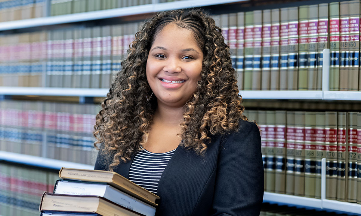 A student smiles while holding a stack of law books in front of shelved volumes in the campus law library, wearing a black blazer over a striped top.