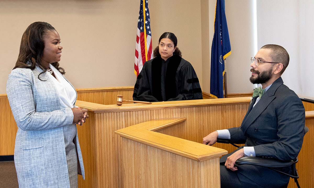 A law student in a blue tweed blazer questions a witness on the stand while a student playing the role of judge presides from the bench during a courtroom simulation exercise.