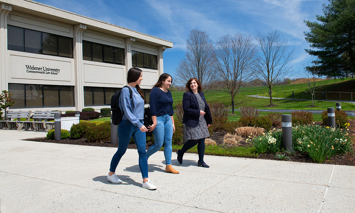 Two students and a staff member walk together along the entrance path of the Widener University Commonwealth Law School building on a sunny spring day, with daffodils blooming in the foreground.