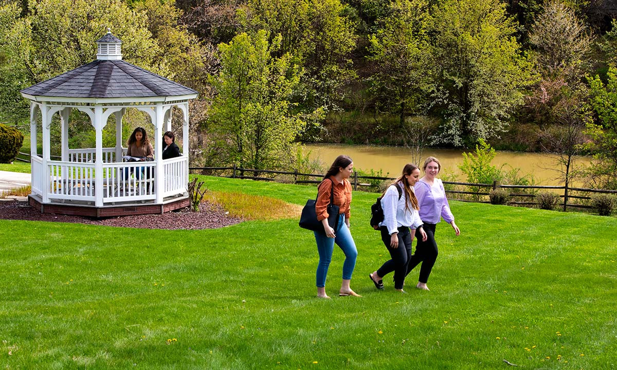Three students walk across a green lawn on the Widener Law Commonwealth campus, with two other students studying in a white gazebo beside a pond and spring trees in the background.