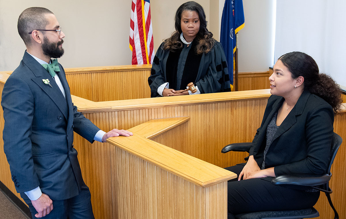 Three students participate in a moot courtroom exercise, with one student presiding as judge in robes and holding a gavel while two others take on the roles of counsel and witness.