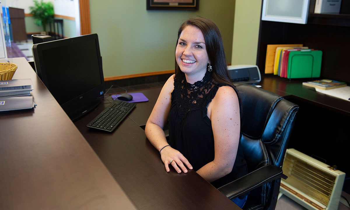 A smiling staff member seated at the admissions reception desk, with a computer, organized files, and office materials visible behind her.