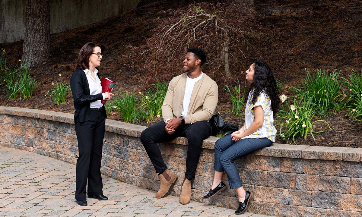 A faculty member converses with two students seated on a stone garden wall outdoors on campus, with spring daffodils blooming in the background.