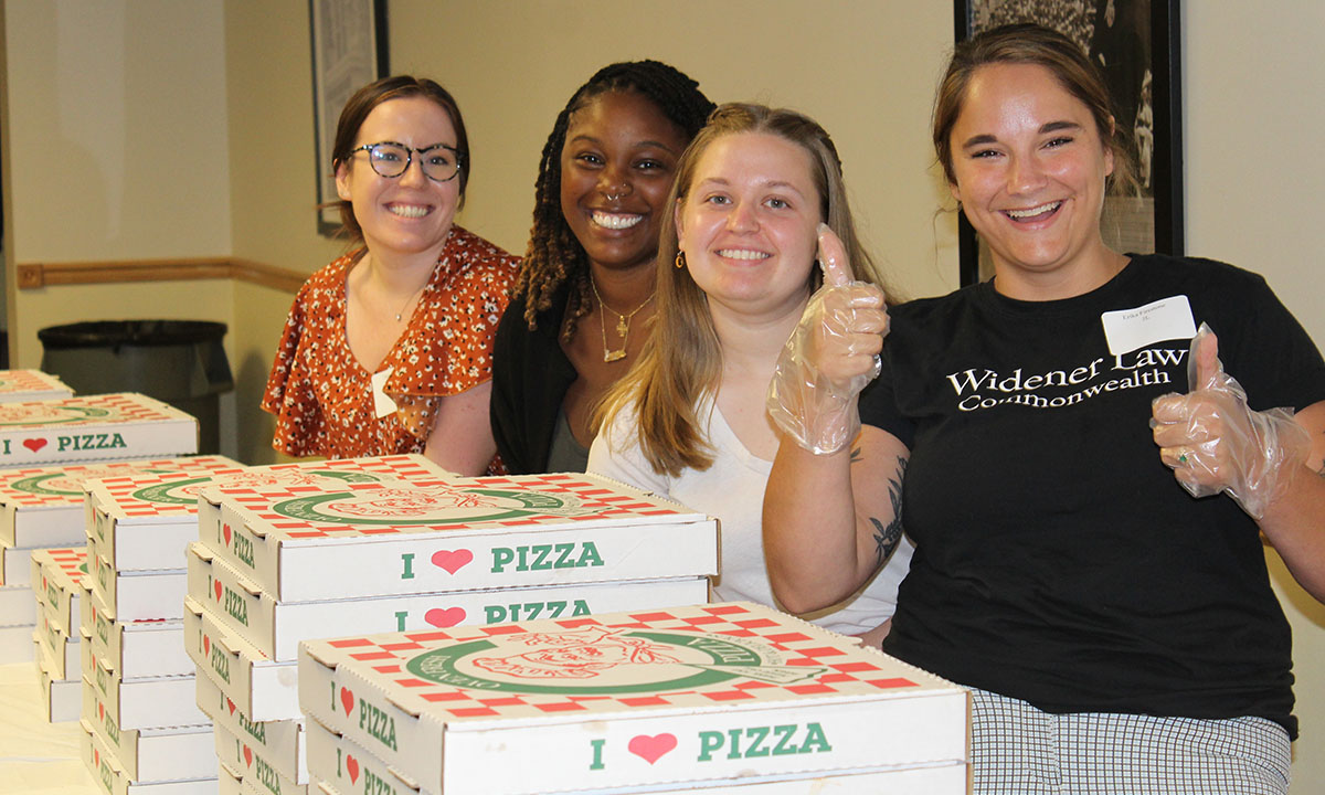 Four Widener Law Commonwealth student ambassadors smile behind stacks of pizza boxes at a campus event, one wearing a Widener Law Commonwealth t-shirt and giving a thumbs up.