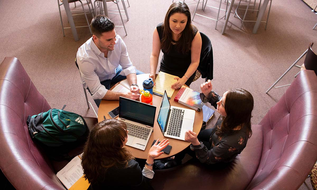Four students collaborate around a round table with laptops and textbooks open, photographed from above in a lounge area with curved seating.