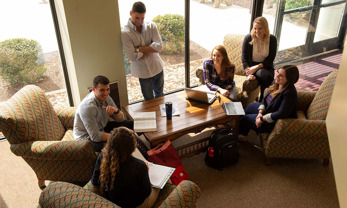 Six students gather around a coffee table in a sunlit lobby lounge area, with laptops and notebooks open, engaged in casual group study or discussion.