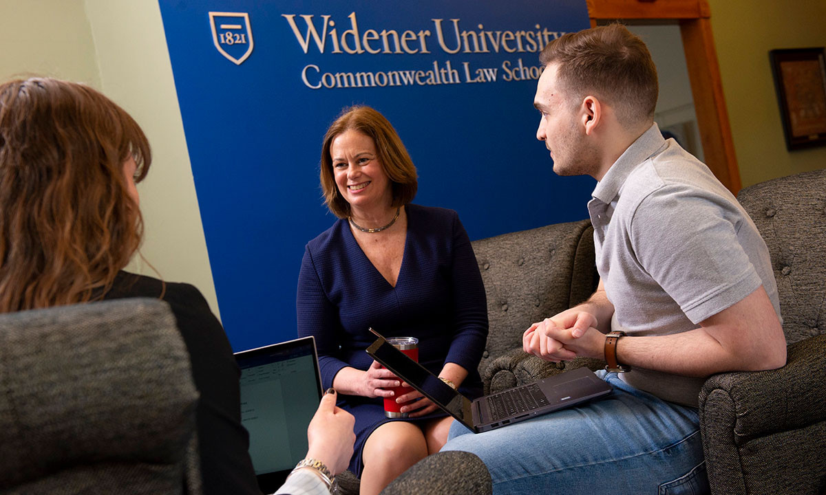 A faculty member in a navy dress meets with two students seated on a couch, laptops open, in front of a Widener University Commonwealth Law School banner.