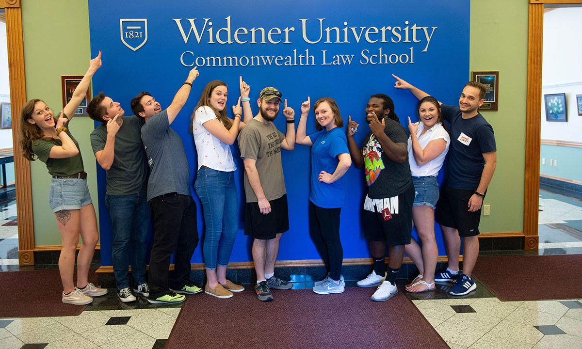 A group of students poses enthusiastically in front of the Widener University Commonwealth Law School lobby sign, many pointing upward at the school name.