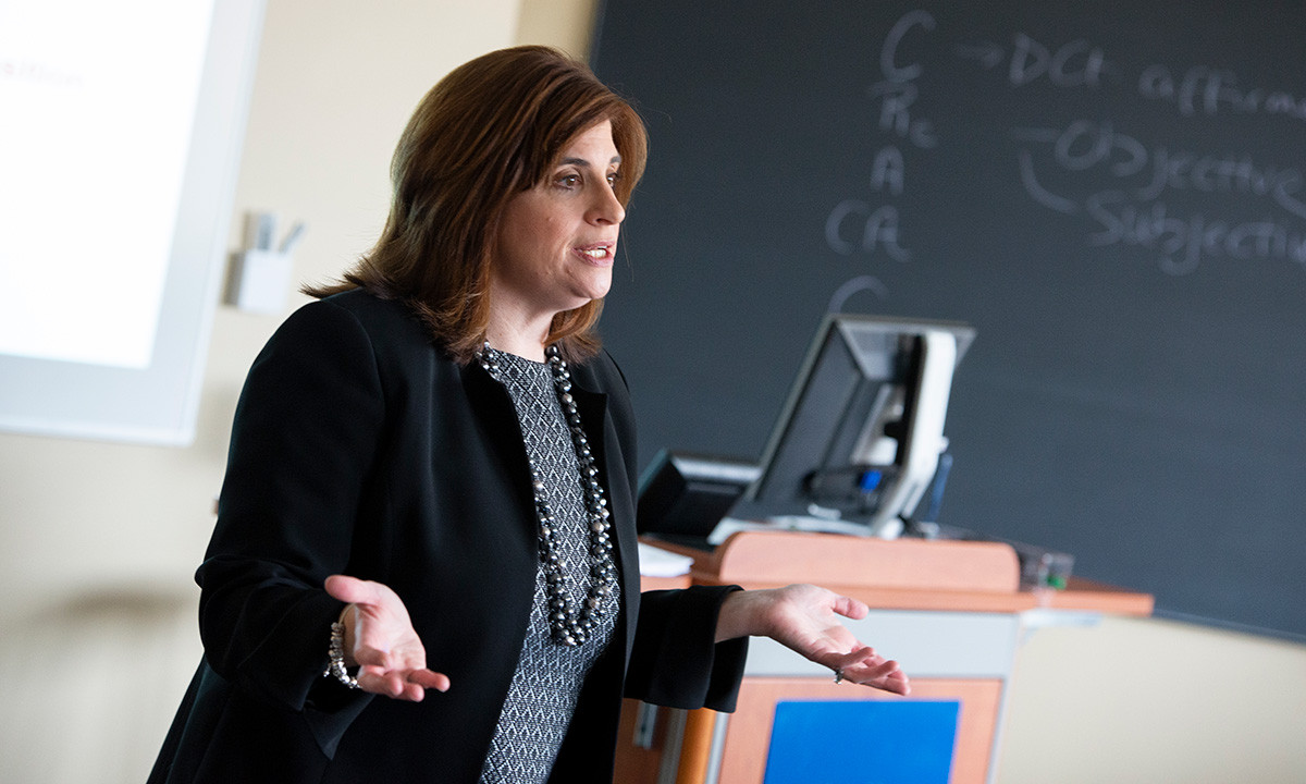 Randi Blackman Teplitz gestures while teaching at the front of a classroom, with legal notes visible on the chalkboard behind her, wearing a black blazer and statement necklace.
