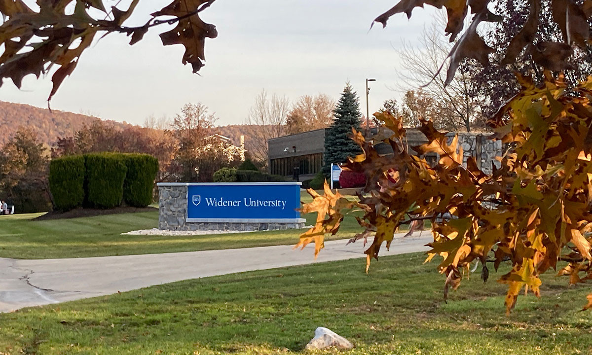 The Widener University campus entrance sign stands along a drive lined with fall foliage, with tree-covered hills in the background.
