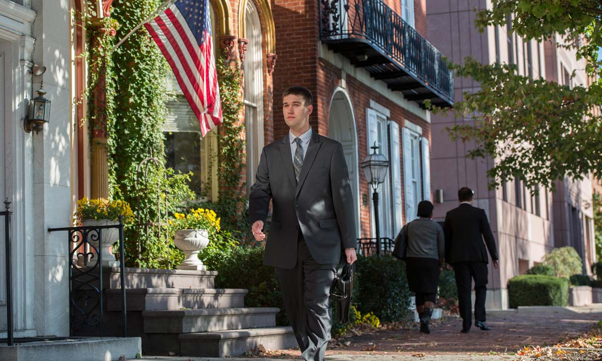 A law student in a gray suit carries a briefcase while walking along a tree-lined brick sidewalk past ivy-covered buildings and an American flag in downtown Harrisburg.