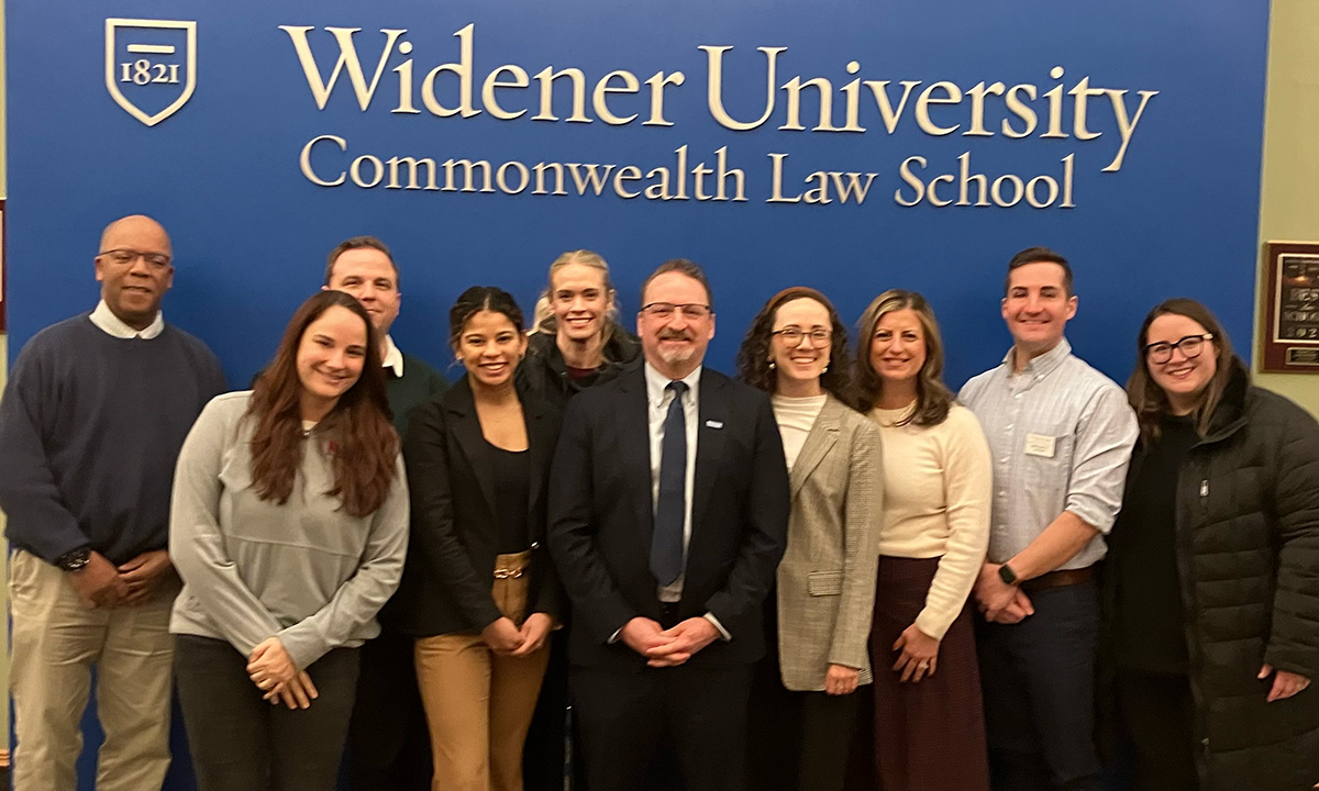 Members of the Widener Law Commonwealth Alumni Advisory Board pose as a group in front of the school's blue lobby sign at their 2025 meeting.