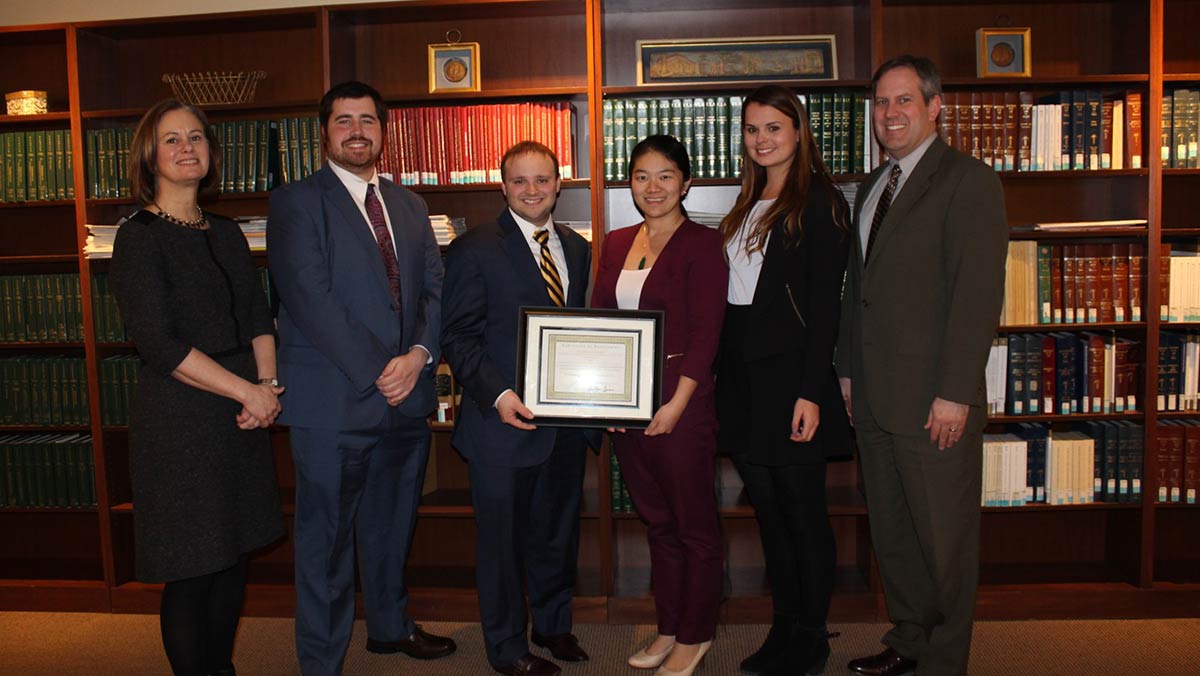 A faculty advisor, three law students, and a coach pose with a framed award certificate in front of law library shelves, celebrating a Business Advising Competition victory.