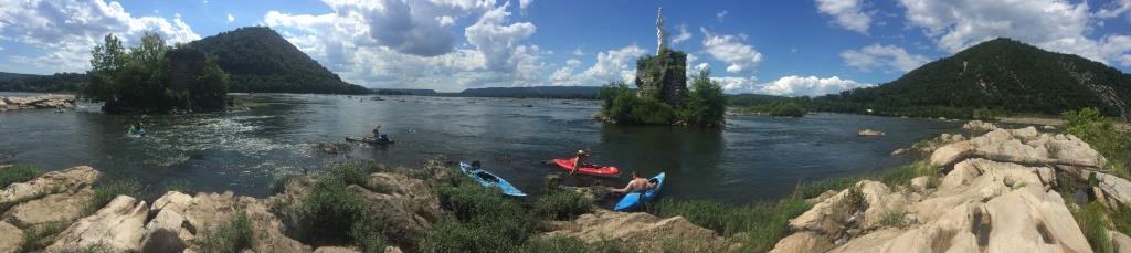 Kayakers rest along a rocky riverbank on a sunny summer day, with tree-covered hills and a stone tower ruin rising from the middle of the river in the background.