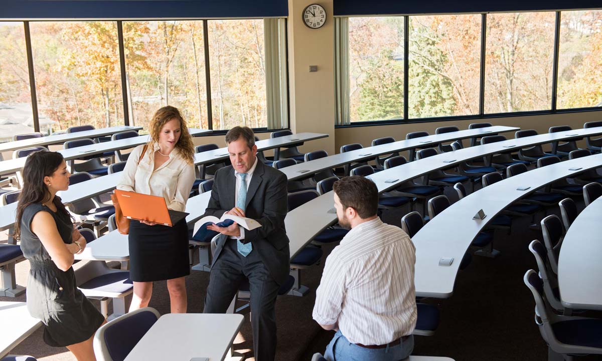 A faculty member consults with two students after class in a tiered lecture hall, with fall foliage visible through the large windows behind them.