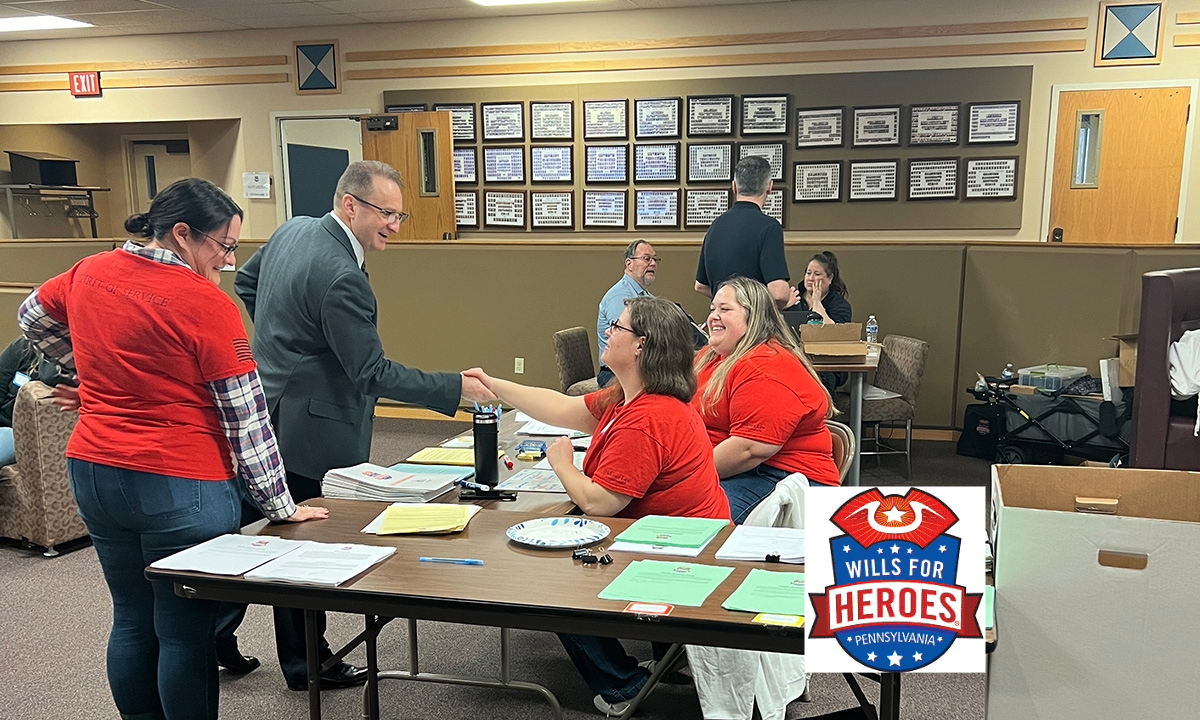 Volunteers in red Wills for Heroes shirts assist participants at document-covered tables during a Wills for Heroes Pennsylvania event, while a man in a suit shakes hands with one of the volunteers. The Wills for Heroes Pennsylvania logo appears in the lower right corner.