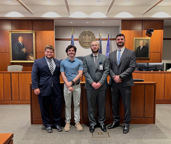 Four students standing in front of a judge's bench with flags in the background.