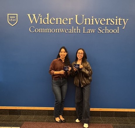 Michelle and Johana standing in front of the blue wall with Widener Law Commonwealth logo behind them. 