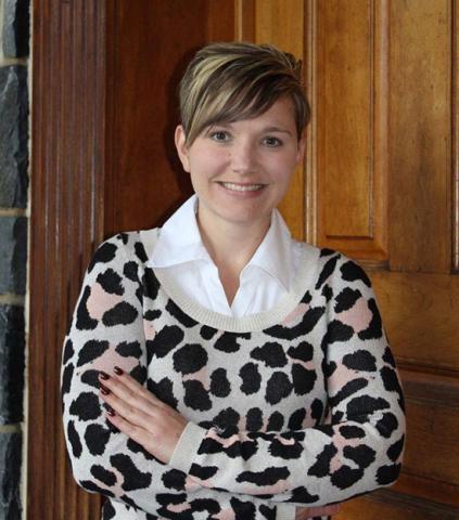Emily Farren smiles with arms crossed in a portrait taken in front of a wooden door, wearing a leopard-print sweater over a white collar shirt.