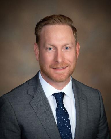 Jake Gilboy poses in a professional headshot wearing a gray suit and navy tie against a warm studio background.