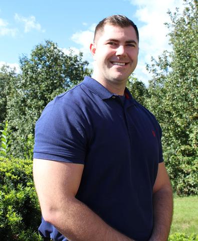 Derek Hartman smiles in an outdoor portrait, wearing a navy polo shirt with trees and blue sky in the background.