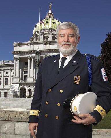 John Logan poses in full dress uniform holding a white cap in front of the Pennsylvania State Capitol building.