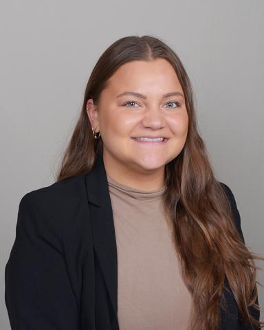 Madelyn Singley: Smiling woman in a black blazer over a taupe turtleneck, with long brown hair, against a neutral gray background.