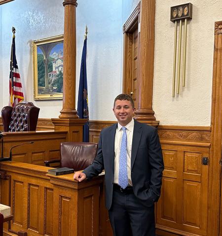 Matthew Strubinger — Matthew Strubinger stands at the podium inside a wood-paneled courtroom at the Carbon County Public Defender's Office, with an American flag and Pennsylvania state flag visible behind him.