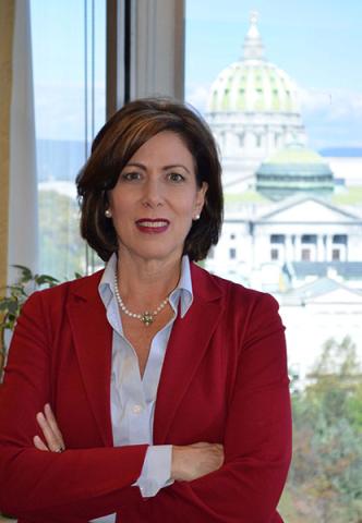 Susan Schwab - Smiling woman in a red blazer with a pearl necklace, arms crossed, with the Pennsylvania State Capitol dome visible through the window behind her.