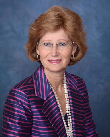 Elizabeth Simcox — Smiling woman in a pink and navy striped blazer with a pearl necklace and rimless glasses, against a blue studio background.