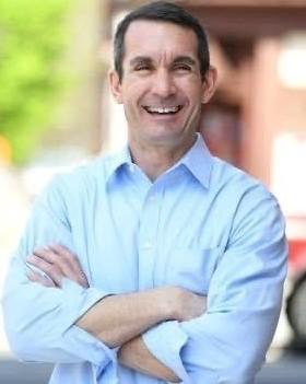 Eugene DePasquale smiles with arms crossed in an outdoor portrait, wearing a light blue shirt.