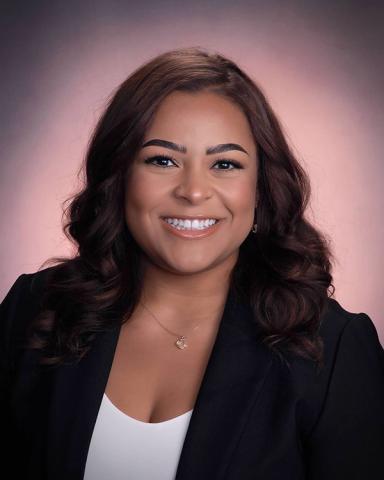 Vanessa Peck: Smiling woman in a black blazer with a delicate gold necklace, against a soft pink gradient background.
