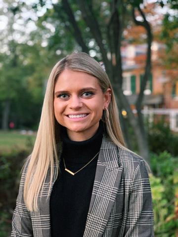 Maranda Moyer — Smiling woman in a plaid blazer and black turtleneck, photographed outdoors with autumn foliage in the background.