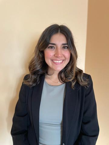 Sarafina Ramalho — Smiling woman in a black blazer over a gray top, against a warm neutral background.