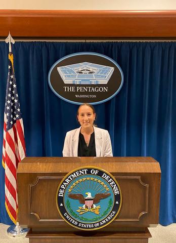 Emily LaGreca — Emily LaGreca stands at the Pentagon press briefing podium beneath the Pentagon seal, with an American flag beside her and a blue curtain backdrop.