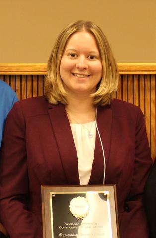 Kayla Rost smiling and holding an award plaque, wearing a burgundy blazer, in a wood-paneled room.