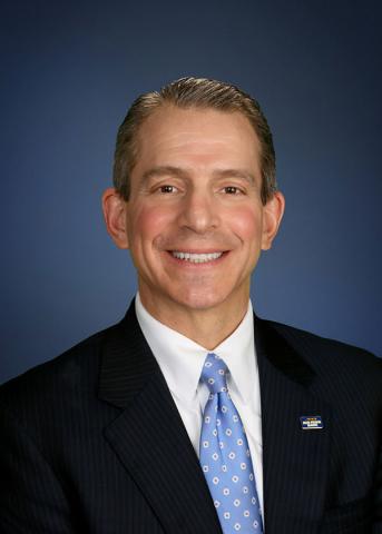 Rory Ritrievi smiles in a professional headshot wearing a dark suit and blue tie against a dark blue background.
