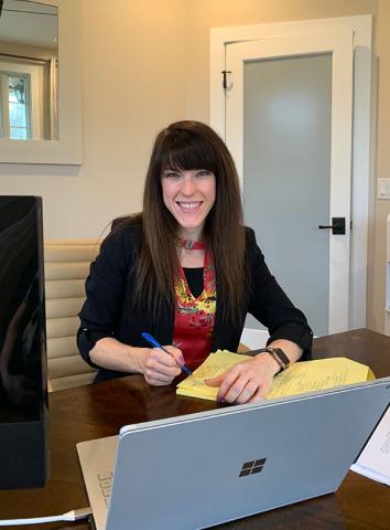 Stephanie Patton smiles while seated at a desk with a notepad and laptop, wearing a black blazer over a floral top.