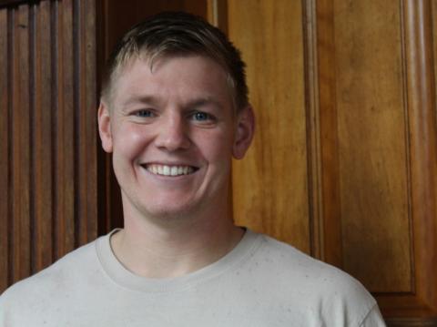 Rob Kranz smiles in a casual portrait in front of a wooden paneled wall, wearing a light gray t-shirt.